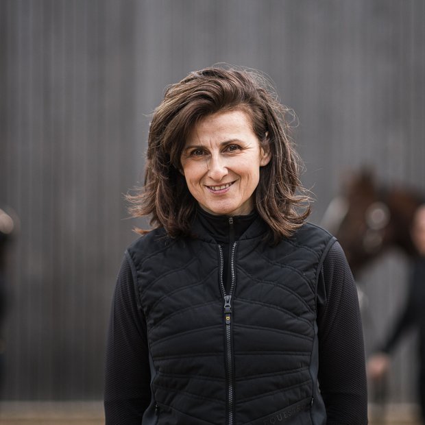 Dove fare equitazione in Alto Adige: hotel con maneggio Donna sorridente con giacca nera davanti a cavalli e persona sfocati