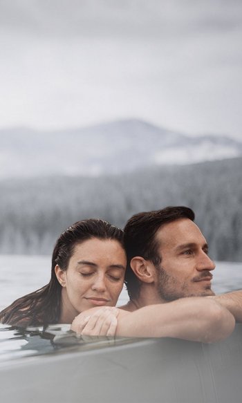 Coppia rilassata in piscina all'aperto con vista sulle montagne in una giornata nuvolosa.