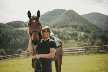 Mann steht mit Pferd auf grünem Feld vor bewaldetem Bergpanorama