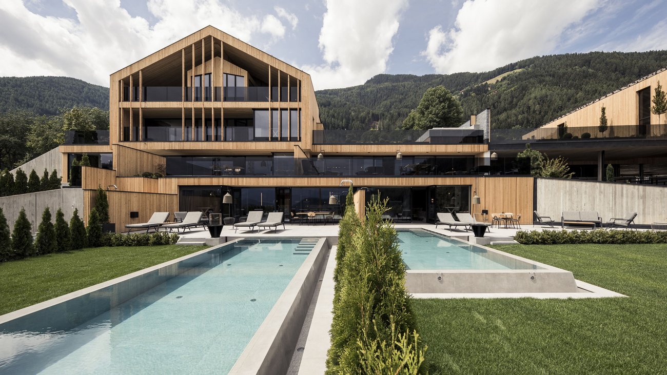 Modernes Holzhaus mit zwei Pools und Liegestühlen vor Bergen unter bewölktem Himmel