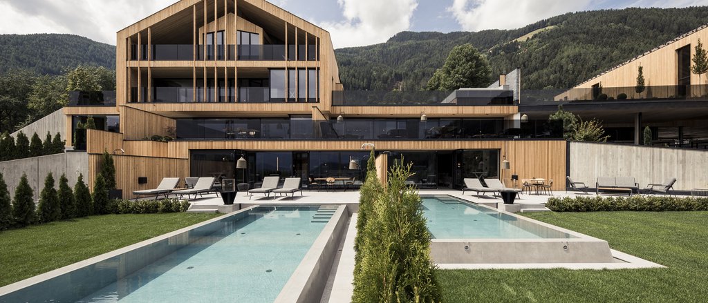 Der facettenreiche Spa unseres Spa-Hotels in Südtirol Modernes Holzhaus mit zwei Pools und Liegestühlen vor Bergen unter bewölktem Himmel