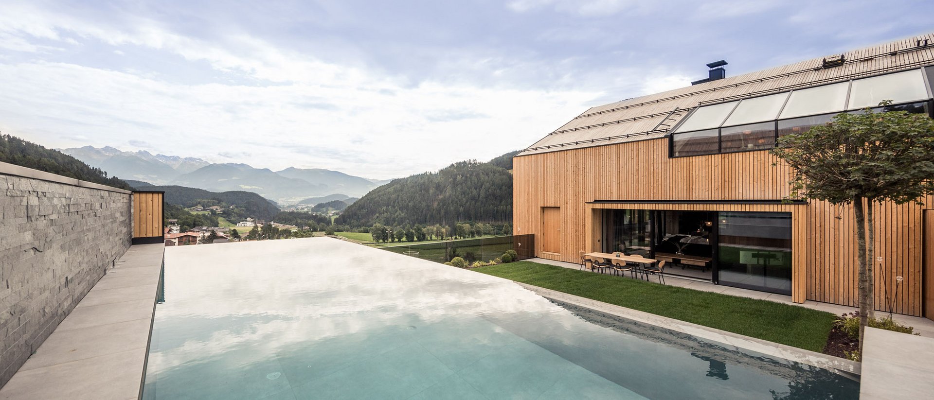 Moderner Holzhaus mit Infinity-Pool und Bergblick bei bewölktem Himmel
