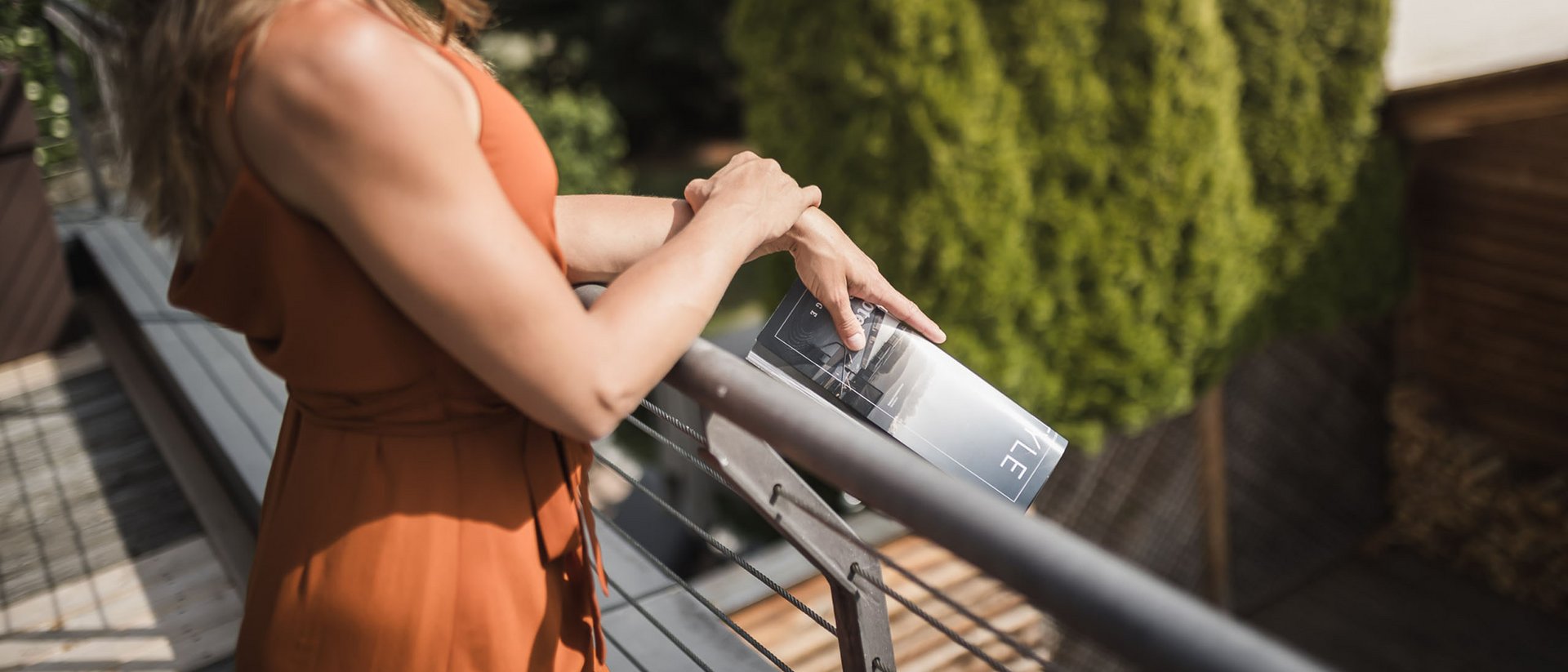 Donna con vestito arancione si appoggia al balcone con una rivista in mano