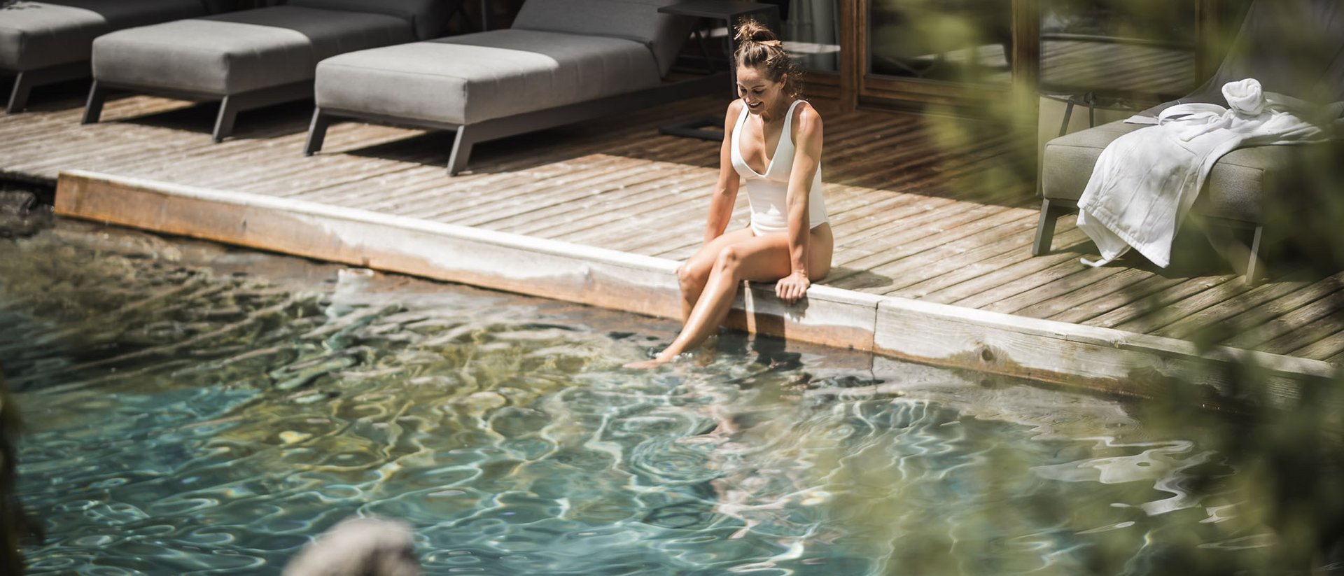 Frau im weißen Badeanzug sitzt am Pool und taucht ihre Füße ins Wasser