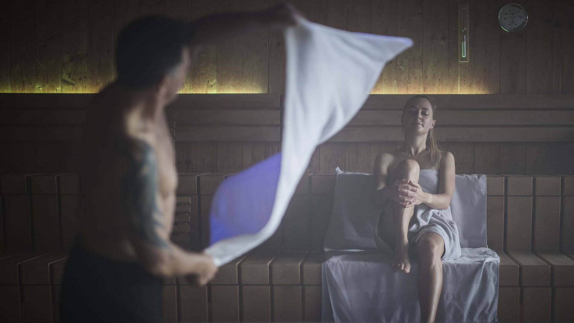 Coppia rilassata nella sauna, uomo agita un asciugamano, donna seduta su panca