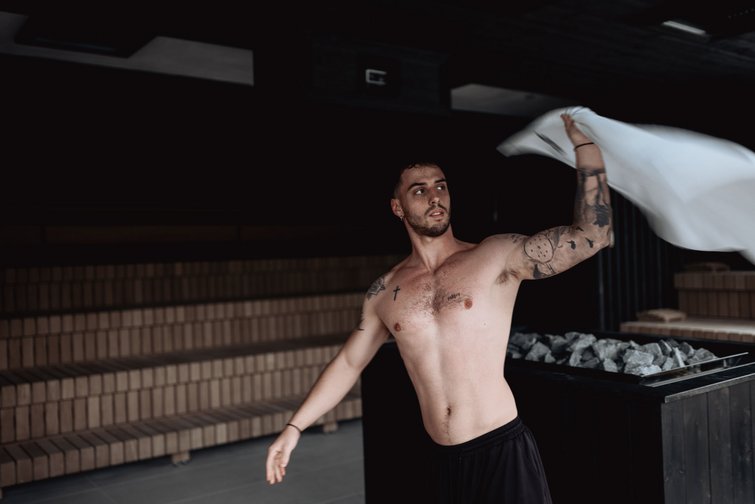 Sauna Week Uomo che agita un asciugamano in sauna con sfondo scuro e fornetto di pietre