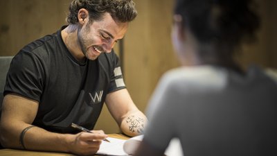 Uomo sorride mentre scrive su carta al tavolo con donna in primo piano