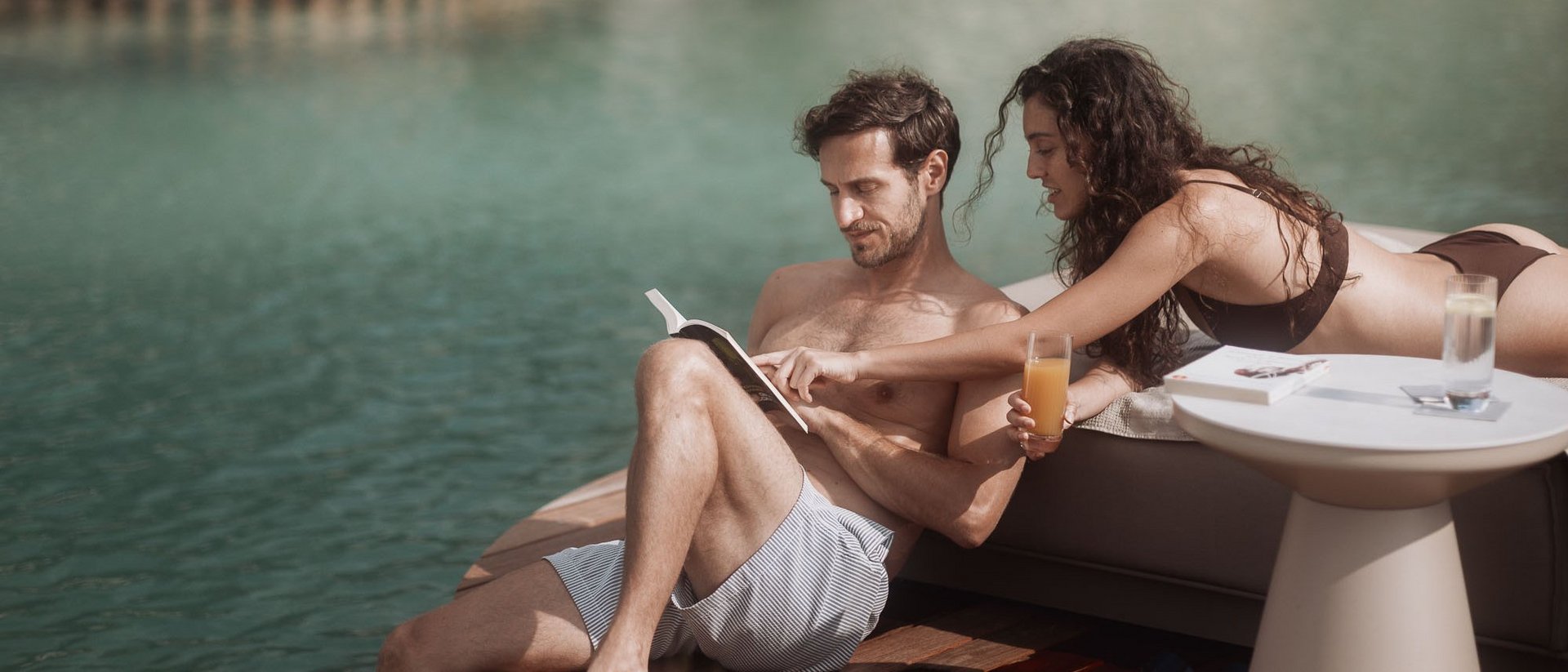 Coppia rilassata a bordo piscina che legge un libro e beve succo