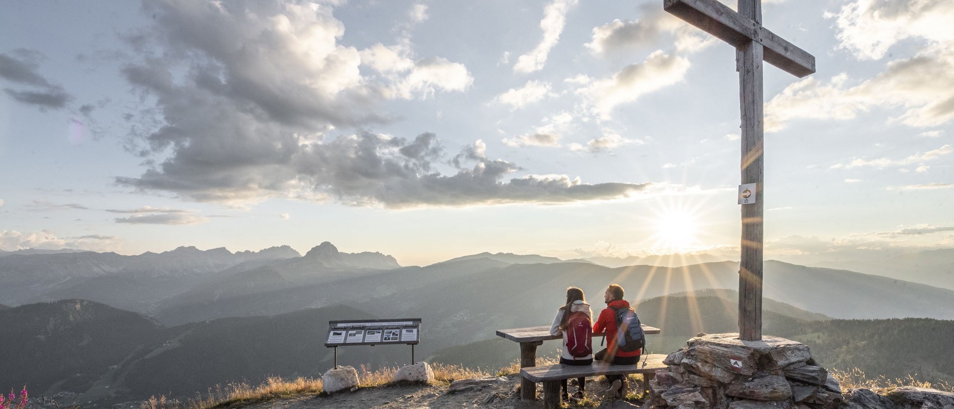 Blick auf Berggipfel mit zwei Wanderern und Holzkreuz bei Sonnenuntergang