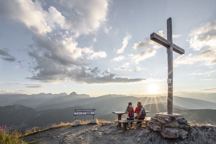 Blick auf Berggipfel mit zwei Wanderern und Holzkreuz bei Sonnenuntergang