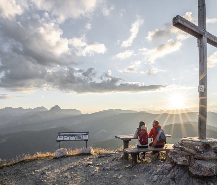 Blick auf Berggipfel mit zwei Wanderern und Holzkreuz bei Sonnenuntergang