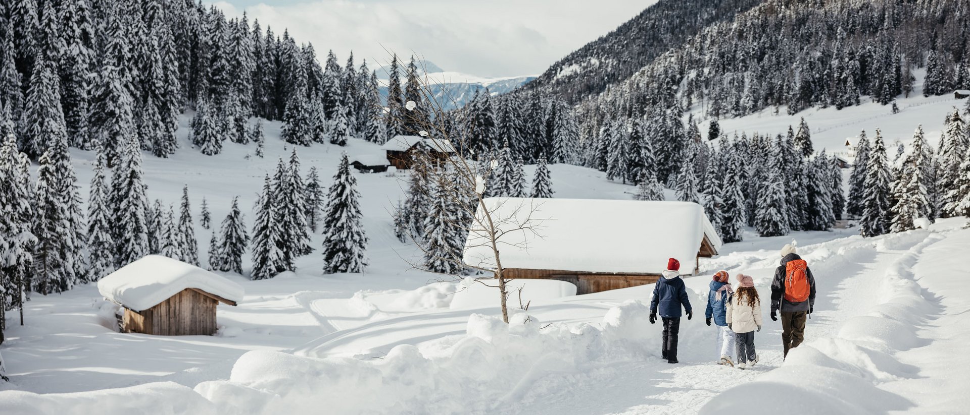 Familie wandert auf verschneitem Weg umgeben von Bergen und Tannenbäumen
