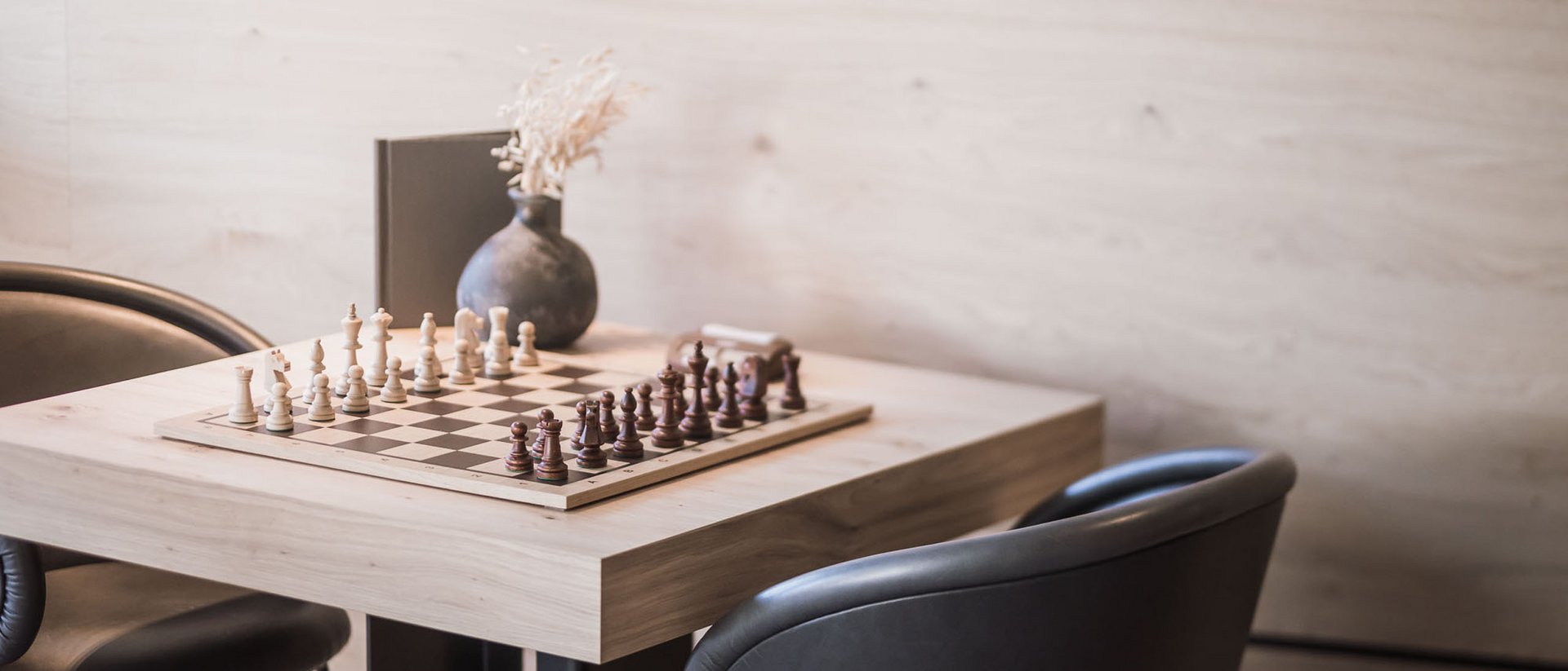 Schachbrett auf Holztisch mit zwei schwarzen Stühlen und einer Vase im Hintergrund