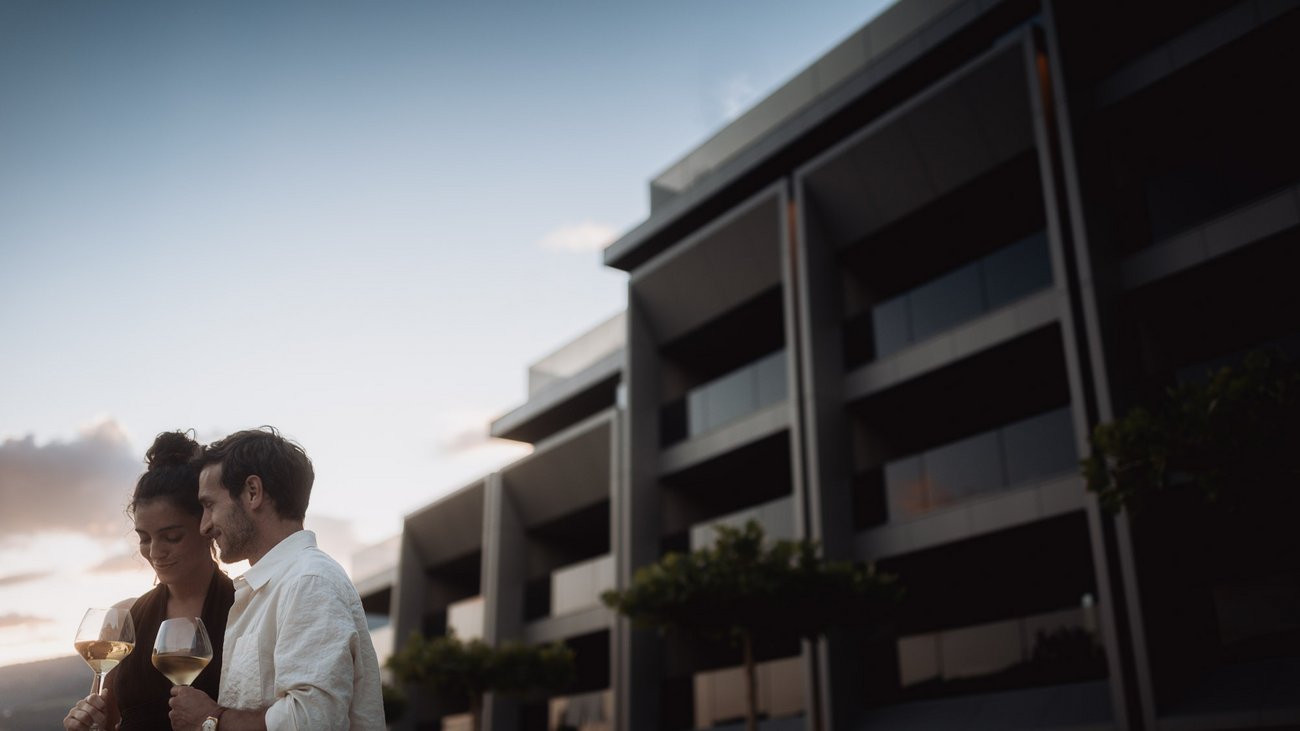 Außergewöhnliche Erlebnisse, Reiten und Yoga im Urlaub Paar genießt Wein auf Balkon vor modernem Gebäude bei Sonnenuntergang