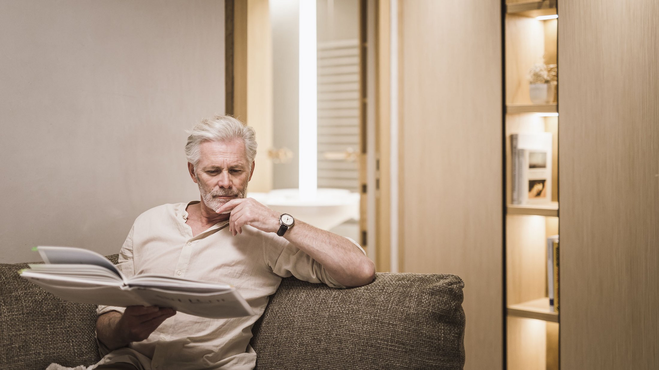 Uomo anziano legge su un divano in una stanza moderna ben illuminata