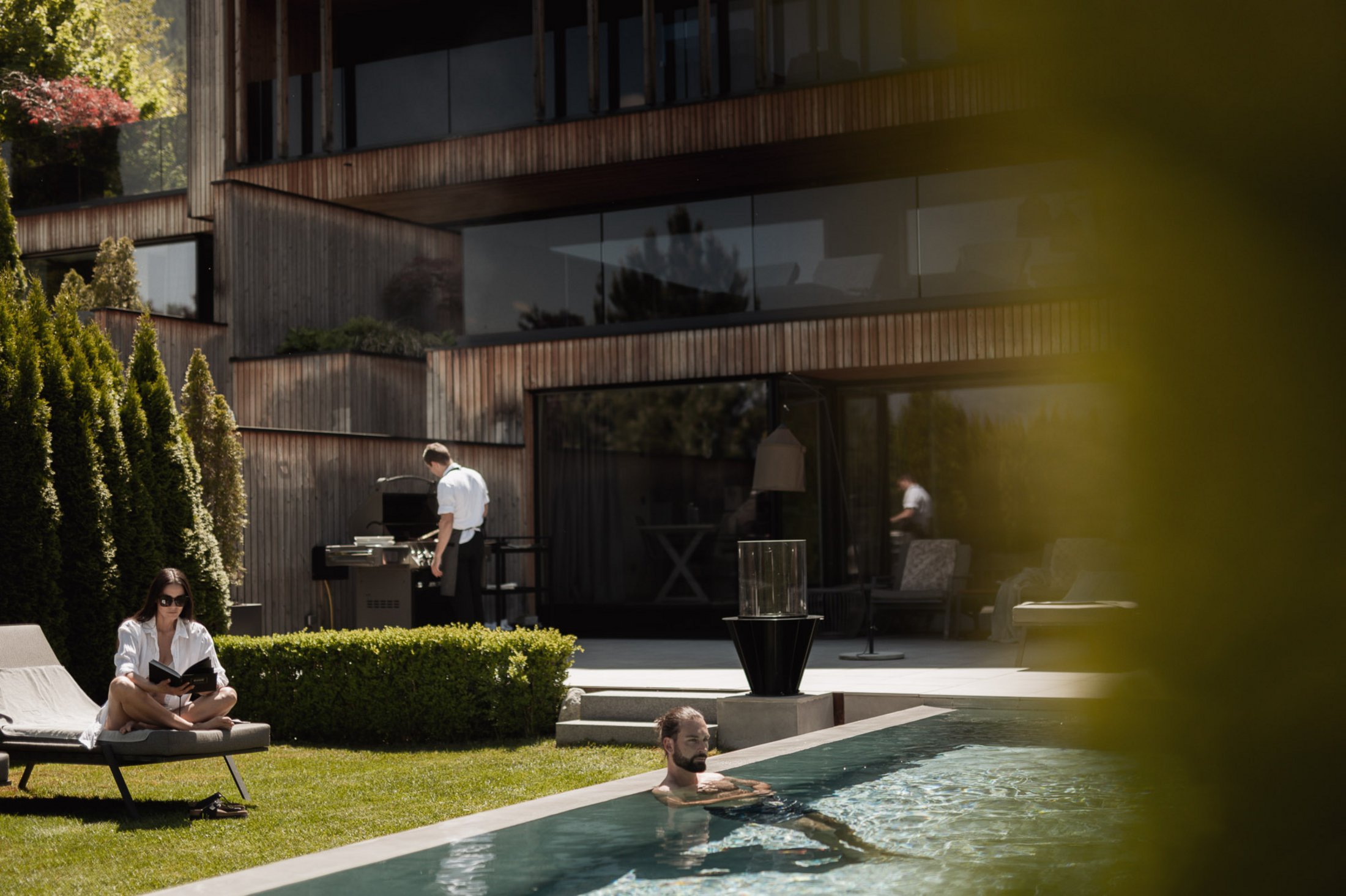 Person reading on lounge chair, man swimming in pool, chef grilling by modern house