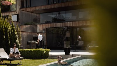 L’esclusività in foto: il nostro chalet in Alto Adige di lusso Persona legge su sedia a sdraio, uomo nuota in piscina, cuoco al barbecue davanti a casa moderna