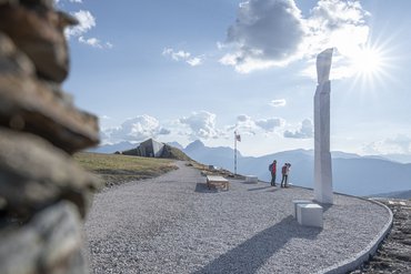 Escursionisti in montagna guardano una scultura con cielo soleggiato e montagne