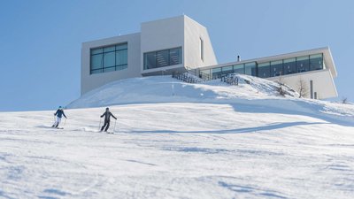 Due sciatori davanti a un edificio moderno su una scogliera innevata con cielo sereno