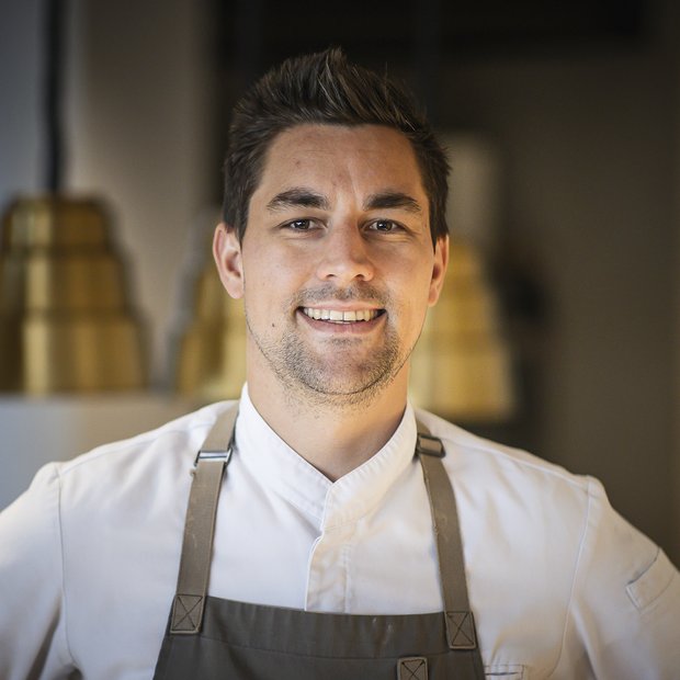 Lächelnder Koch in weißem Hemd und grauer Schürze vor unscharfem Hintergrund