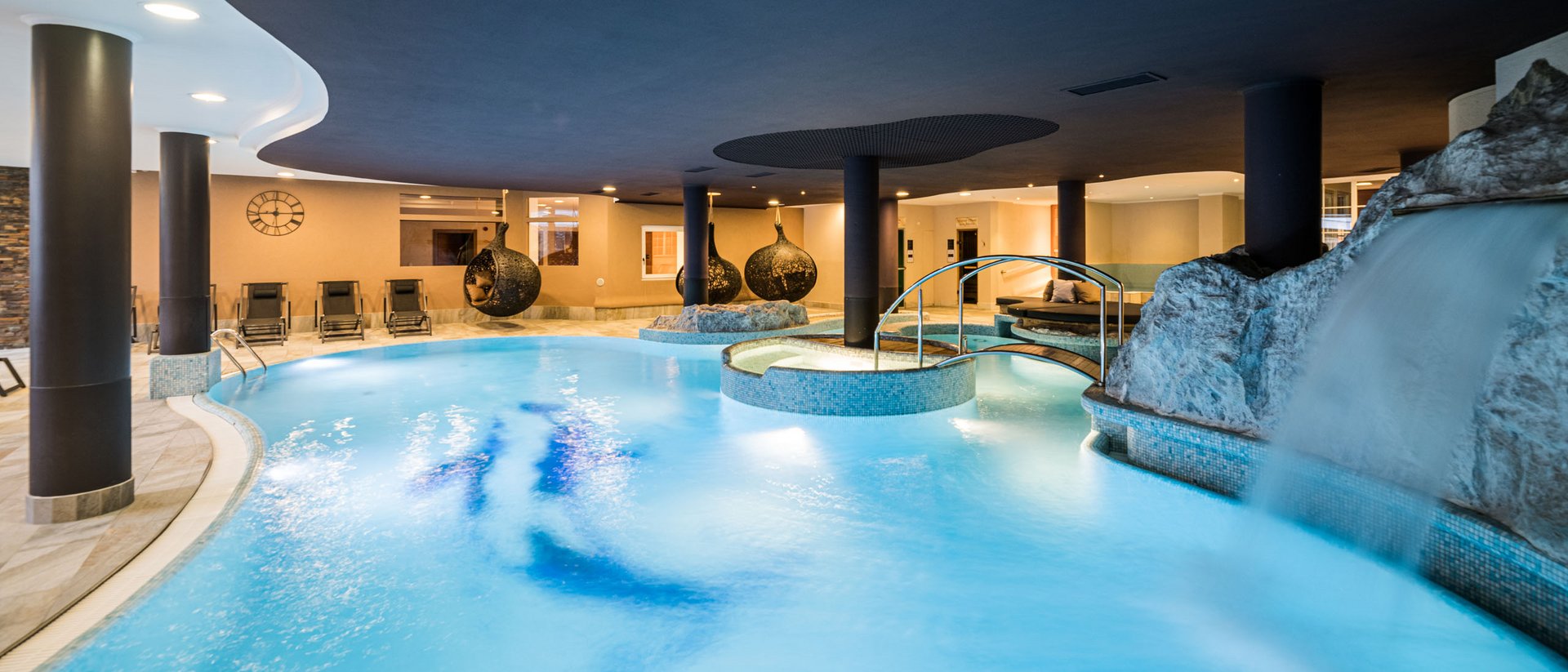 Innenbereich mit beleuchtetem Pool, Wasserfall und Liegestühlen