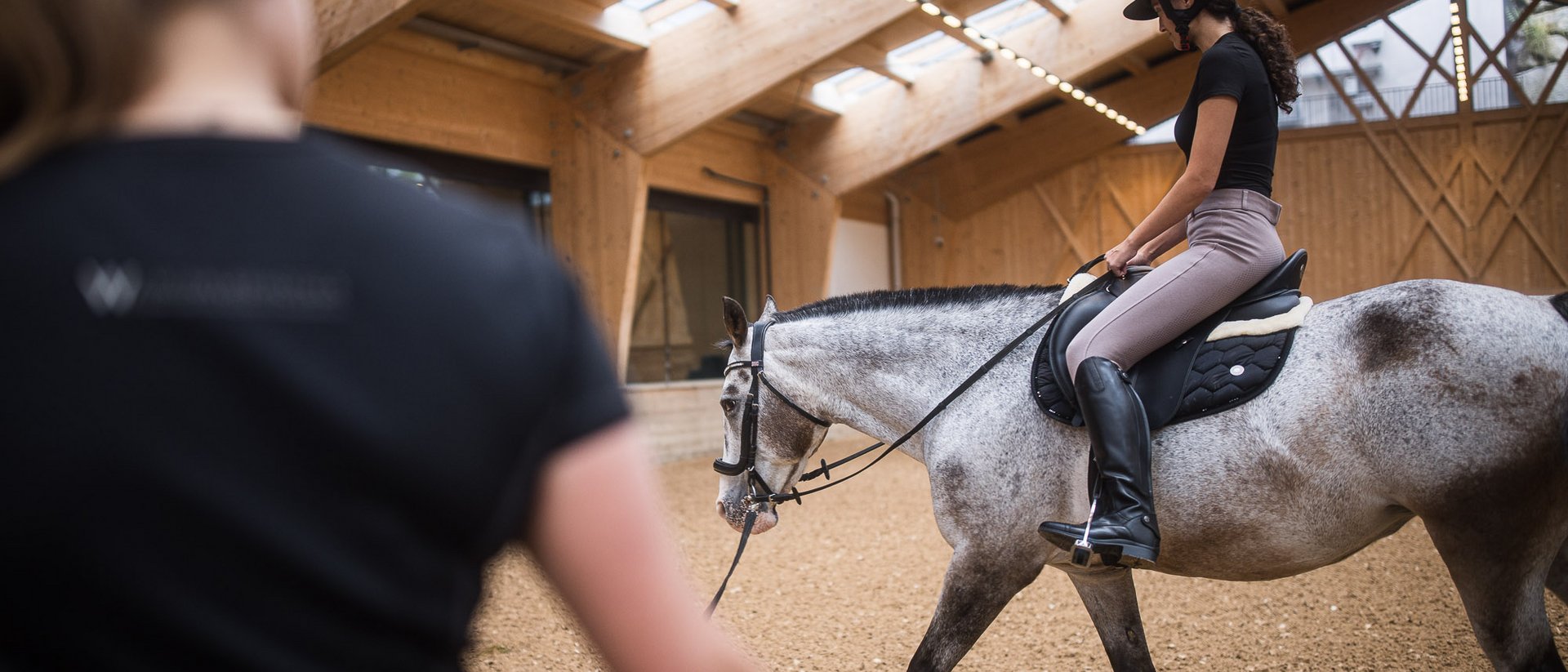 Frau reitet auf Pferd in Reithalle mit Trainer im Vordergrund