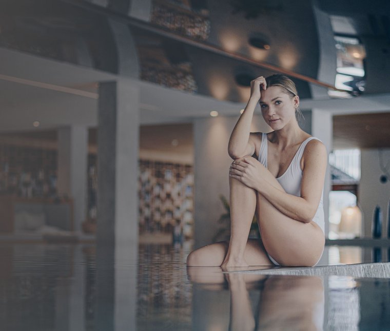 Donna in costume bianco seduta al bordo di una piscina in ambiente interno moderno