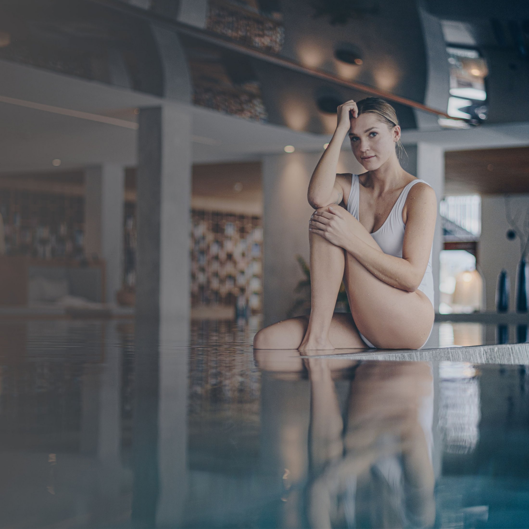 Frau im weißen Badeanzug sitzt am Rand eines Pools in modernem Innenbereich