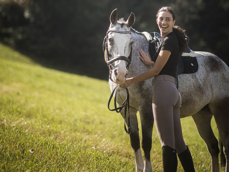 Woman in riding attire smiling beside saddled horse in a grassy field