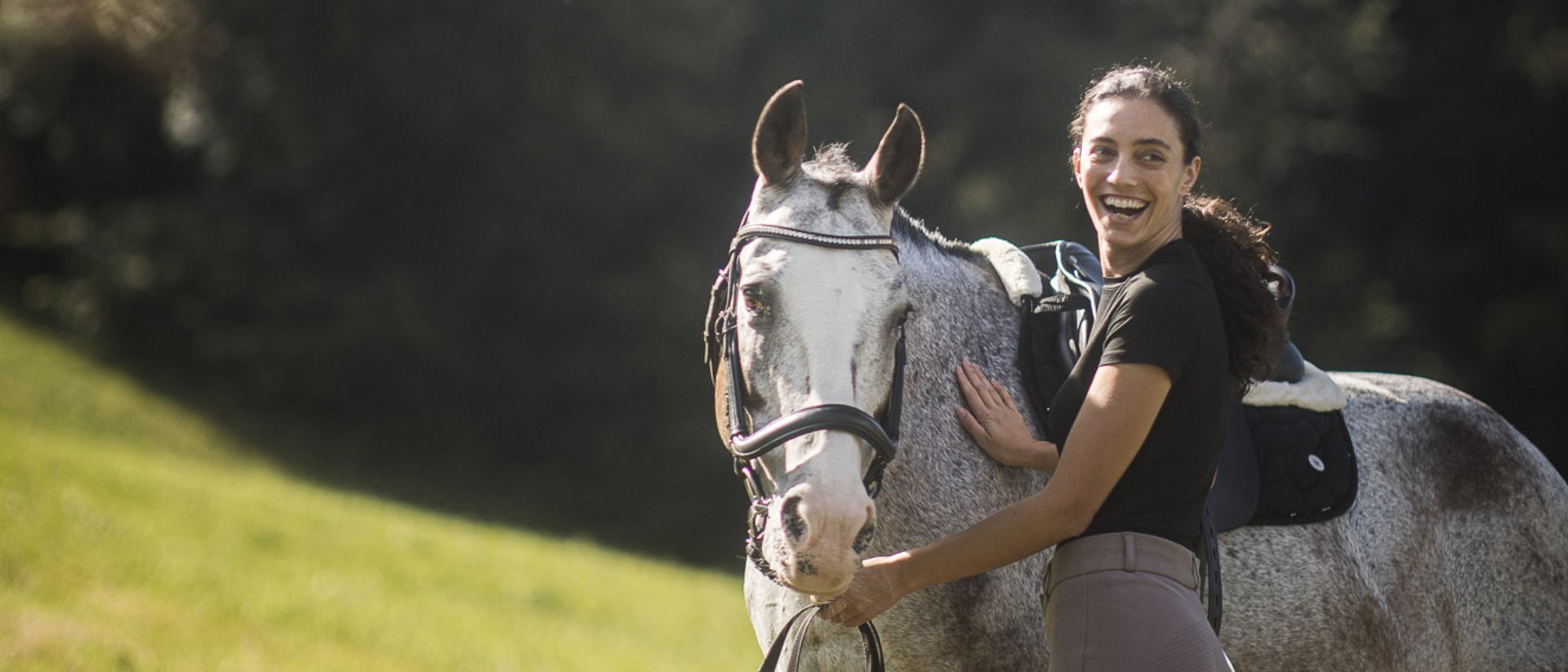 Dove fare equitazione in Alto Adige: hotel con maneggio Donna in abbigliamento da equitazione sorridente accanto al cavallo sellato in un prato