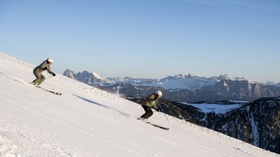 Due sciatori scendono una pista innevata con cielo sereno e montagne sullo sfondo