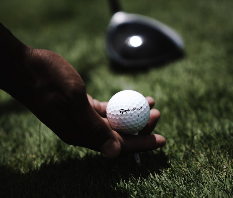 Hand legt Golfball auf Abschlag auf grünem Gras mit Golfschläger im Hintergrund