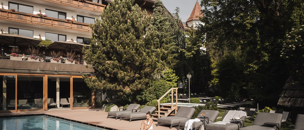 Der facettenreiche Spa unseres Spa-Hotels in Südtirol Frau sitzt am Pool vor Hotel mit Liegestühlen und Bäumen