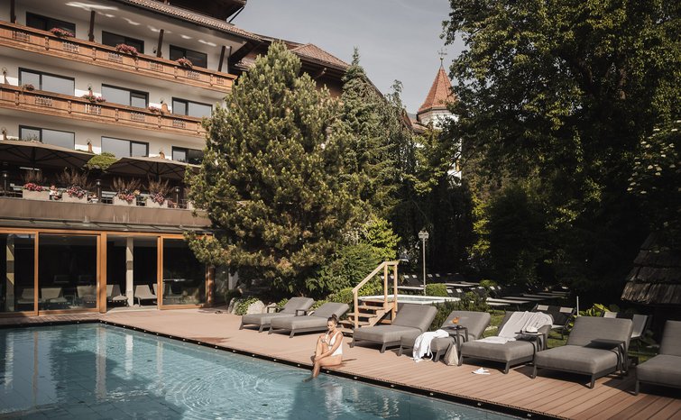 Woman sitting by pool in front of hotel with lounge chairs and trees
