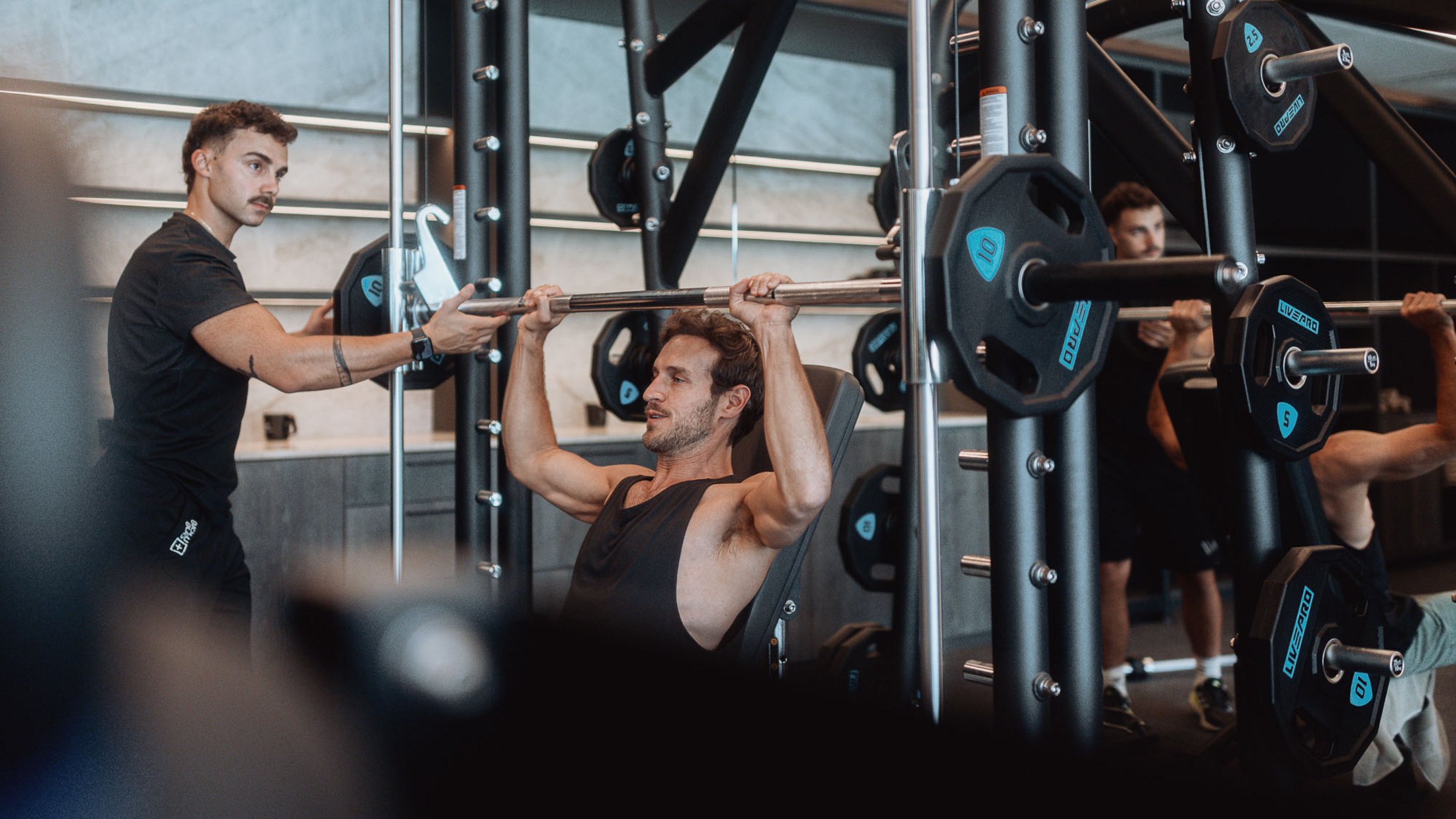 Mann trainiert Schulterdrücken mit Langhantel im Fitnessstudio, Trainer unterstützt
