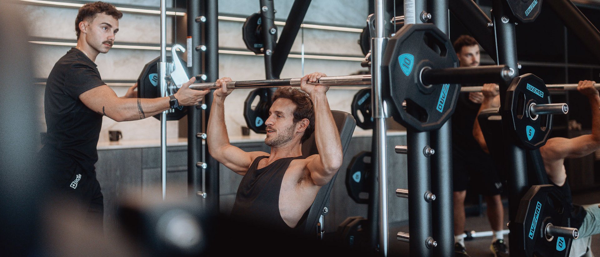 Yoga und Fitness in Ihrem Sporthotel in Südtirol Mann trainiert Schulterdrücken mit Langhantel im Fitnessstudio, Trainer unterstützt