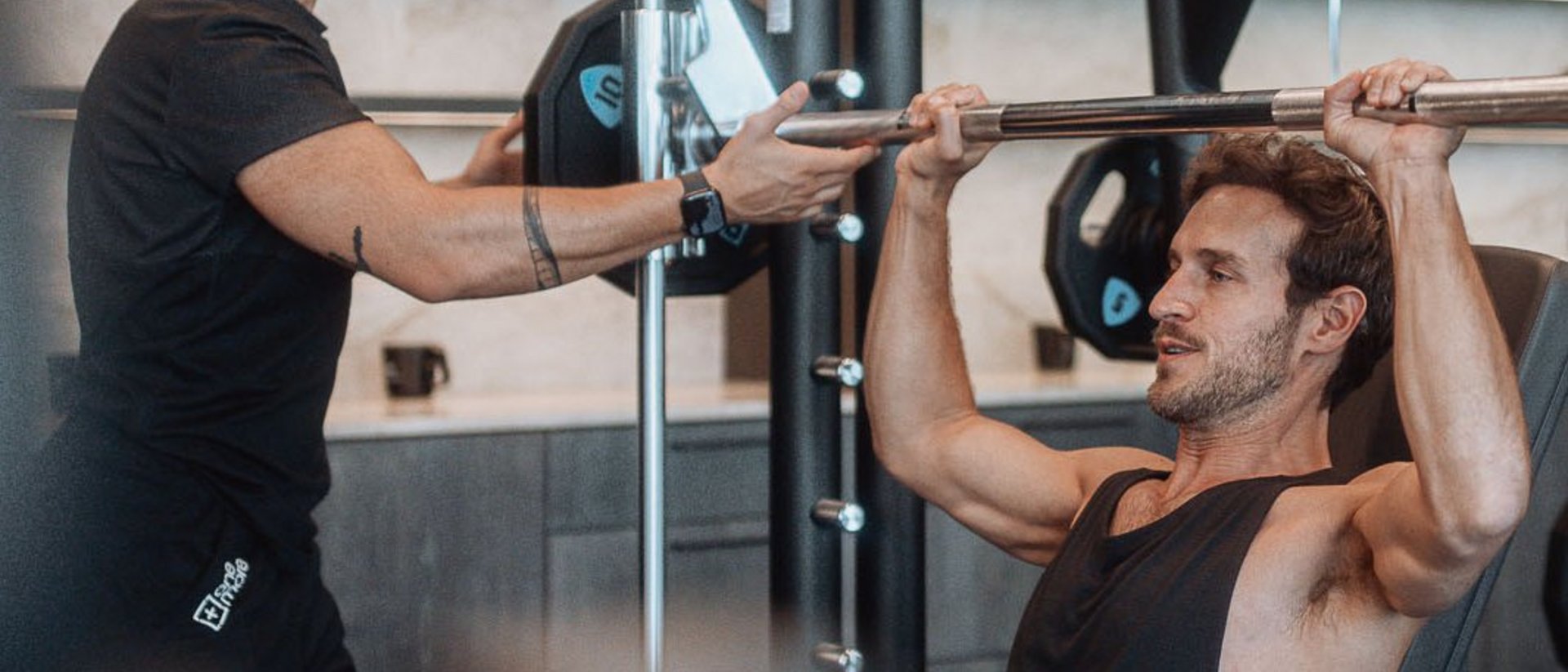 Yoga und Fitness in Ihrem Sporthotel in Südtirol Mann trainiert Schulterdrücken mit Langhantel im Fitnessstudio, Trainer unterstützt