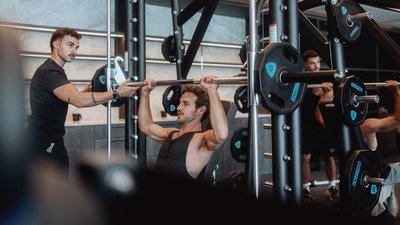 Photos from your hotel in Pfalzen in Val Pusteria/Pustertal Man doing shoulder press with barbell in gym, trainer assisting him