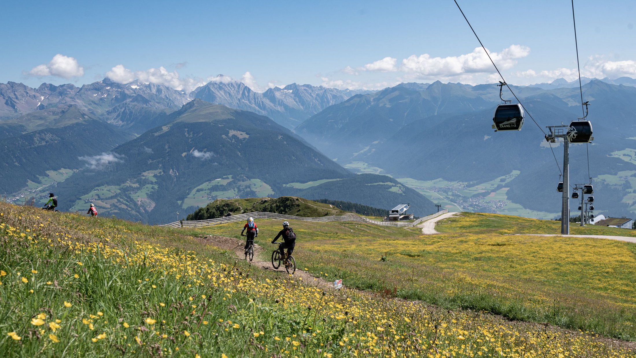 Mountainbiker fahren auf blumiger Wiese mit Bergblick und Gondelbahn