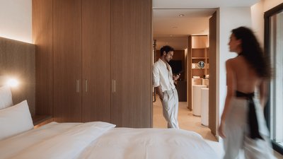 Photos from your hotel in Pfalzen in Val Pusteria/Pustertal Man leaning on wall in hotel room, woman walking past, bed in foreground