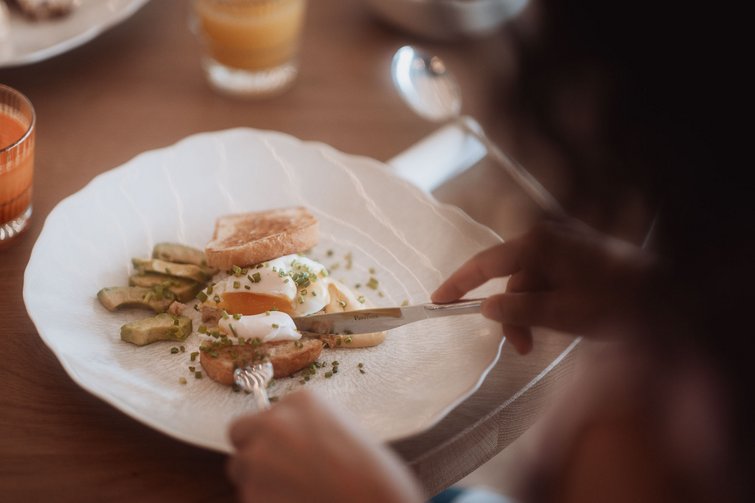 Person schneidet ein pochiertes Ei mit Toast und Avocado auf einem weißen Teller