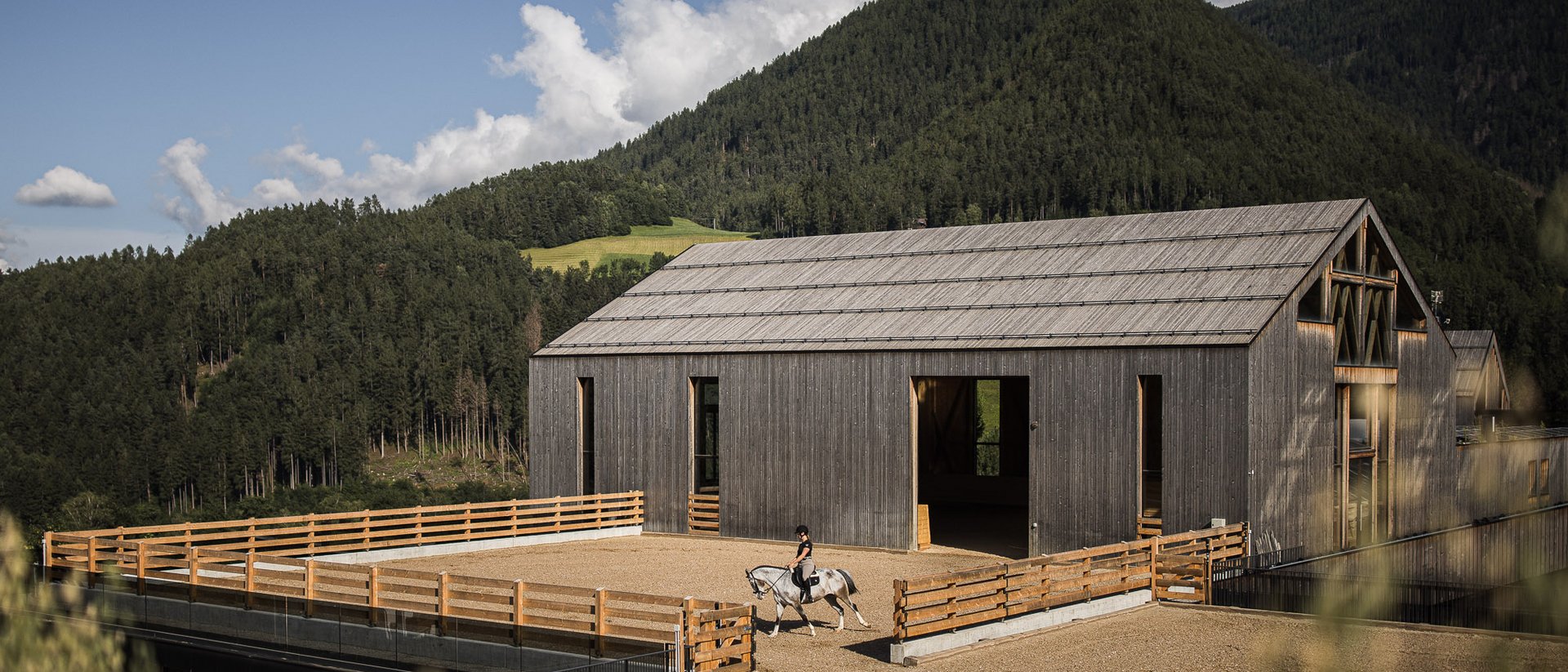 Reiter auf Pferd vor modernem Holzstall in Berglandschaft