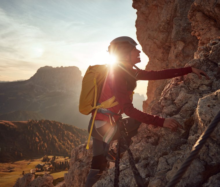 Frau klettert mit Helm und Rucksack an felsiger Bergwand bei Sonnenuntergang