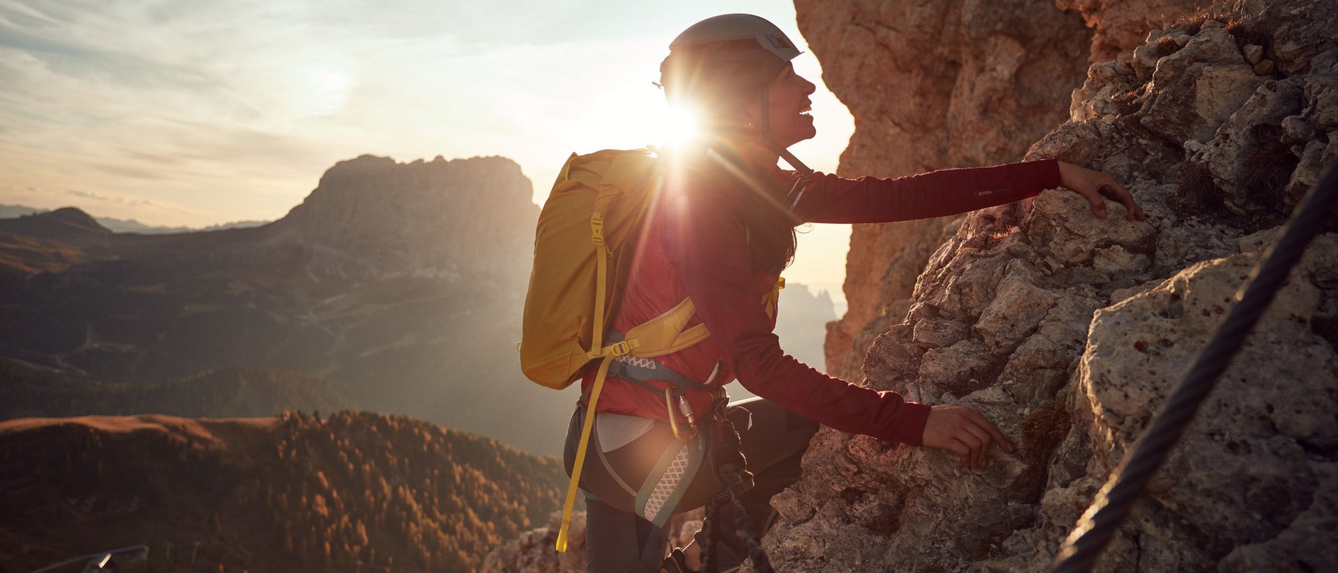 Frau klettert mit Helm und Rucksack an felsiger Bergwand bei Sonnenuntergang