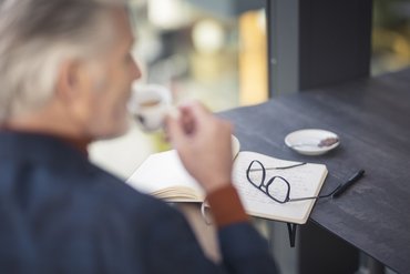 Older man drinking coffee, notebook with glasses and pen on the table