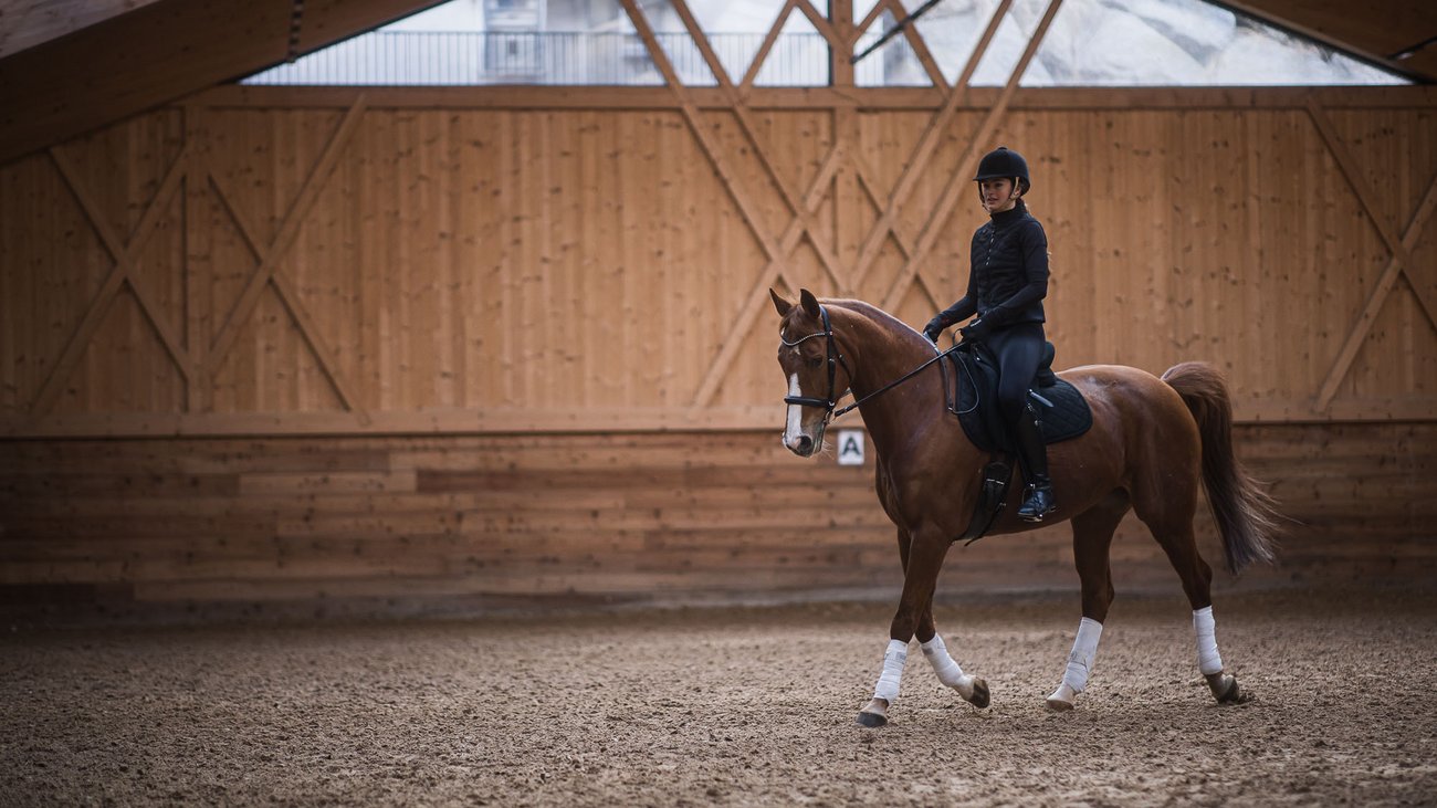 Außergewöhnliche Erlebnisse, Reiten und Yoga im Urlaub Reiterin auf braunem Pferd in Reithalle mit Holzverkleidung