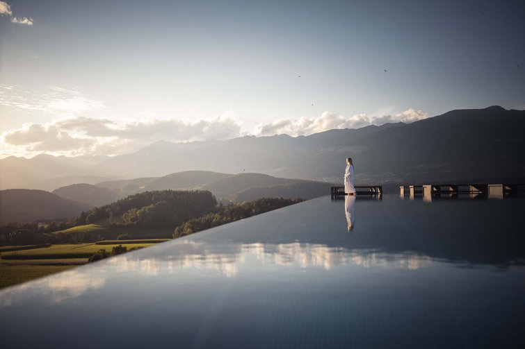 Woman in bathrobe standing at pool edge overlooking mountain landscape