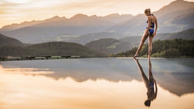 Donna in costume da bagno blu sul bordo di una piscina a sfioro con vista sulle montagne al tramonto