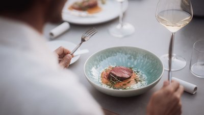 Person holding fork over plate with gourmet dish and glass of white wine