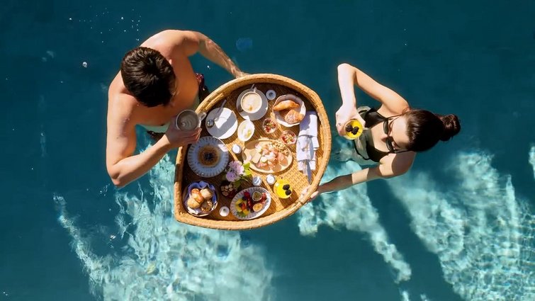 Man and woman in pool with floating breakfast basket full of food and drinks